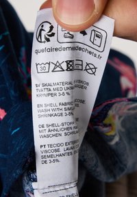 Label tag met verzorgingsinstructies in meerdere talen op een donkere, gemustreerde stoffen achtergrond. Materialen zijn onder andere viscose met een krimpwaarschuwing van 3-5%.