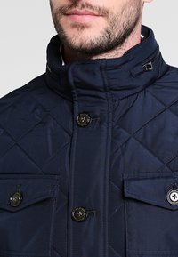 Marinblå vadderad jacka med hög krage, två framfickor och stora svarta knappar. Har en dragkedja och stickad diamantmönster.