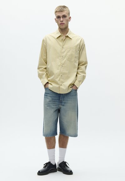 Keltainen raidallinen nappipaita, farkkushortsit, mustat kengät ja valkoiset sukat. Rentoon tyyliin; paidassa on tasku, shortsit polvenpituudet.