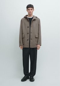 Cappotto di lana marrone con cappuccio, bottoni frontali, due tasche applicate e vestibilità morbida, abbinato a pantaloni e scarpe nere.