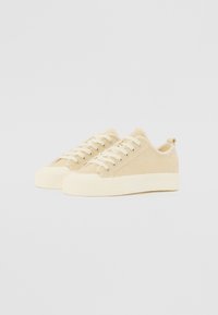 Sneakers in tela beige con una superficie liscia, punta rotonda e suola in gomma piatta. Presentano lacci e un anello sul tallone per una facile vestibilità.
