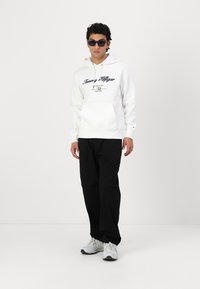 Hvit hettegenser med "Tommy Hilfiger"-logo, kenguru-lomme og snorhette. Kombinert med svarte bukser og grå sneakers.