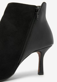 Stivale alla caviglia con tacco alto nero, realizzato in una combinazione di camoscio e pelle liscia. Chiusura con zip laterale, punta affusolata e tacco leggermente conico.