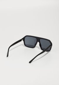 Occhiali da sole neri, oversize, con lenti esagonali, finitura opaca, montatura spessa e aste nere sottili. Le lenti sono di un grigio scuro e piatte.