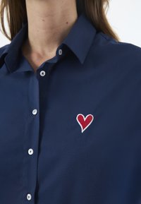 Tmavě modrá košile s knoflíky vyrobená z hladké tkaniny, s červeným vyšitým srdcem na levé straně a klasickým designem límečku.