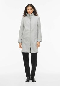 Pelēks flīsa mētelis ar augstu apkakli, priekšējo r�āvējslēdzēju, sānu kabatām un gariem piedurknēm, kombinēts ar melnām biksēm un potīšu zābakiem.