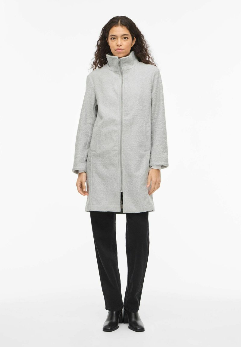 Pelēks flīsa mētelis ar augstu apkakli, priekšējo rāvējslēdzēju, sānu kabatām un gariem piedurknēm, kombinēts ar melnām biksēm un potīšu zābakiem.