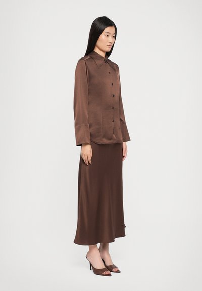 Brown satin long-sleeve shirt with large collar and button-down front, paired with a matching ankle-length skirt and open-toe brown mules.