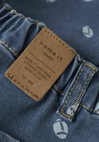 Tkanina dżinsowa w kolorze indygo z subtelną fakturą, z brązową skórzaną metką z napisem "name it" oraz informacjami o pielęgnacji. Złote przeszycia.