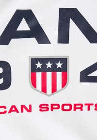 Tissu blanc avec un texte bleu marine audacieux, des accents rouges et un logo en forme de bouclier avec des étoiles et des rayures. Le design comprend les mots « AMERICAN SPORTS ».