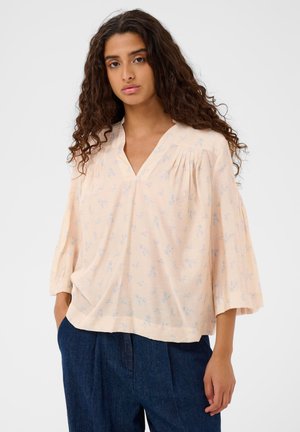 Jeune femme aux longs cheveux bouclés portant une blouse ample couleur pêche pâle avec un petit motif floral et des manches larges, associée à un pantalon bleu foncé.