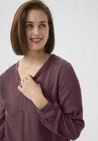 Halenka s výstřihem do V a dlouhými rukávy v tmavě mauve barvě, s texturovanou látkou, jemnými plisováními na ramenou a knoflíkovými manžetami.