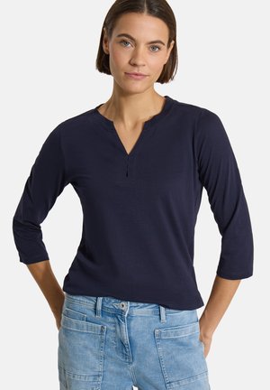 Femme aux cheveux courts et bruns portant un haut bleu marine à manches trois-quarts et un jean bleu clair, les mains posées sur les hanches.