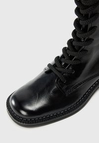 Bottine en cuir noir avec un dessus texturé, bout rond et épais lacets noirs. Semelle en caoutchouc avec des coutures détaillées le long du bord.