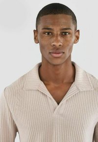 Lys beige ribbet polo-skjorte med tradisjonell krage og halv knappestolpe. Stoffet virker mykt med en subtil tekstur og mønster.