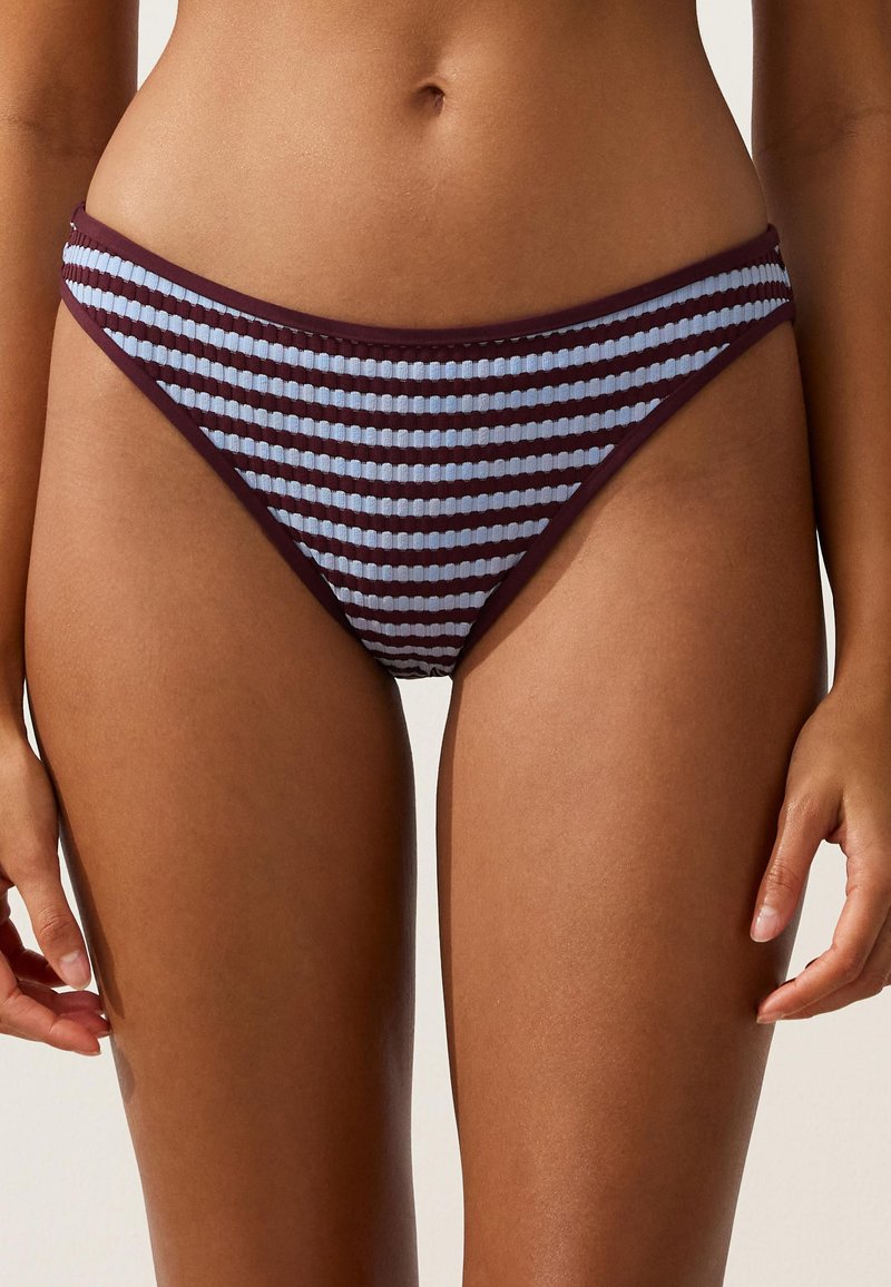 Femme portant un bas de bikini taille haute avec des rayures horizontales bleu clair et violet foncé, montrée du ventre jusqu'aux cuisses supérieures.