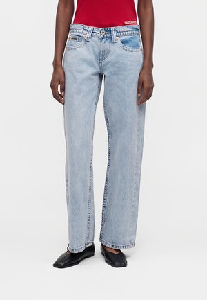 Osoba nosząca jasnoniebieskie jeansy o prostych nogawkach, czarne baleriny z kwadratowym noskiem oraz czerwony bezrękawnik wpuszczony w spodnie.