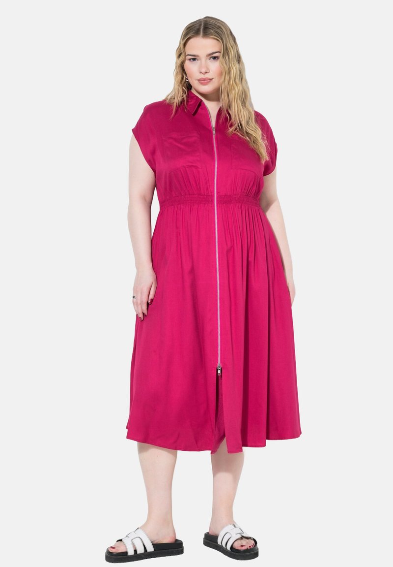 Abito fucsia con maniche corte, chiusura frontale con zip, vita elastica e gonna fluida. Indossato con sandali neri con cinturini bianchi.