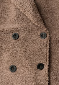 Manteau en polaire marron avec un design croisé, doté de boutons noirs et d'un tissu doux et texturé. Détail de col remarquable.