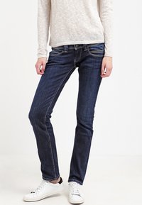 Jeans ajustados de un azul oscuro con detalles de costura visibles, bolsillos estándar y una cintura a media altura, combinados con zapatillas blancas.