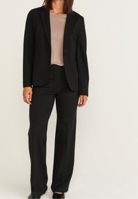 Schwarzer maßgeschneiderter Blazer mit passender Hose, der eine glatte Textur und ein strukturiertes Design aufweist; getragen über einem hellbeigen Oberteil.
