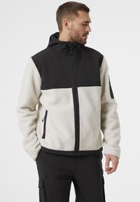 Chaqueta de forro polar con panel superior negro y cuerpo crema, tejido texturizado, cierre de cremallera en el frente y capucha ajustable con cordones.