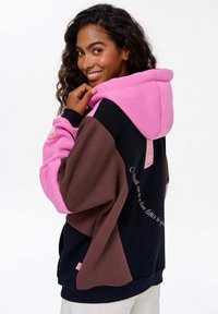 Oversized hoodie w kolorowych blokach w odcieniach różu, brązu i czerni. Posiada kaptur z troczkami oraz nadrukowany tekst na plecach.