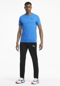 Blauwe katoenen t-shirt met een zwart logo, gecombineerd met zwarte broek met een wit logo en witte schoenen met rode en blauwe accenten.