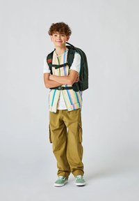 Ein Junge trägt eine mehrfarbige gestreifte Weste über einem weißen Hemd, Cargo-Hosen und grüne Turnschuhe. Er trägt einen dunkelgrünen Rucksack.