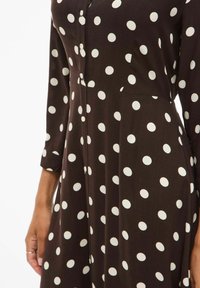 Vestido marrón con grandes lunares blancos, detalle de botones en la parte delantera y mangas largas con puños. Textura de tela suave.