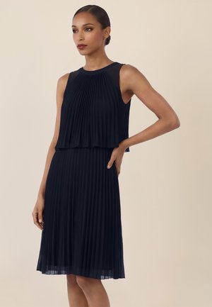 Kobieta w bezrękawniku, plisowanej czarnej sukience, stojąca z jedną ręką na biodrze na tle jednolitego beżowego tła.