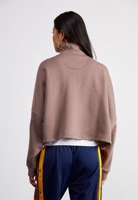 Sweatshirt castanho, cropped, com um alto colar, de corte relaxado e com detalhes cosidos, emparelhado com calças azul-marinho com riscas amarelas laterais.