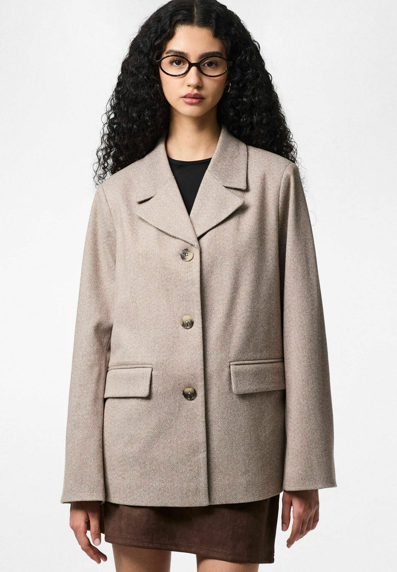 Blazer beige en tissu tissé avec un col cranté, trois gros boutons, deux poches avant et des manches longues. Associé à un haut foncé et une jupe marron.