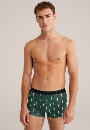 Giovane uomo in piedi senza camisa, che indossa boxer verdi con motivo di facce di mucca bianche e nere, su uno sfondo semplice.
