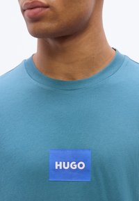 Κοντινό πλάνο ενός ατόμου που φοράει μπλουζάκι σε τιρκουάζ απόχρωση με ένα μπλε ορθογώνιο μπαλώμα στο στήθος, όπου αναγράφεται η λέξη "HUGO" με λευκά γράμματα.