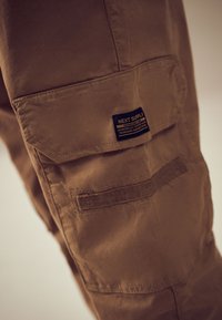 Tasca di pantaloni cargo marroni con patta e etichetta nera con la scritta "NEXT SUPPLY", con dettagli di chiusura in Velcro sul lato della gamba.