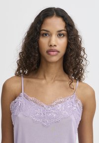 Lavandas krūšturis ar mežģīņu apdari ap kaklu. Tam ir plānas spageti siksnas un maiga, gluda auduma tekstūra.