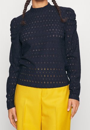 Nainen, jolla on musta, teksturoitu neulepusero, jossa on liehuvat olkapäät, ja kirkkaan keltaiset korkeavyötäröiset housut.