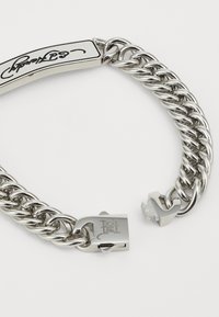 Bracelet en chaîne en métal argenté avec une large plaque ovale d'identification et une signature gravée, comportant un fermoir sécurisé et une finition polie.
