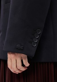 Blazer marine en tissu texturé, avec cinq boutons noirs sur les manches et un logo brodé. Main partiellement visible sur un tissu ressemblant à des plis.