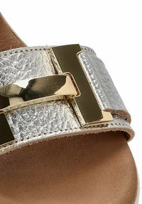 Sandalia de feta plateada metálica con un upper texturizado, detalle de herrajes en tono dorado y una plantilla suave de color beige.