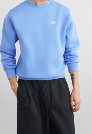 Person iført en lyseblå Nike sweatshirt med ærmerne skubbet op og løse sorte bukser, som viser en tatovering på venstre underarm.