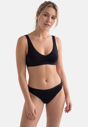 Giovane donna che indossa un reggiseno nero e mutandine nere abbinate, in piedi contro uno sfondo bianco semplice, che guarda leggermente di lato.