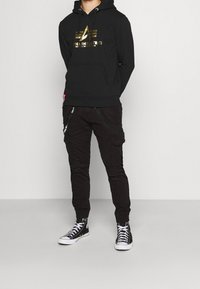 Felpa nera con logo dorato, tasca a marsupio, abbinata a pantaloni cargo neri con tasche laterali e cerniere, indossati con sneakers alte nere.