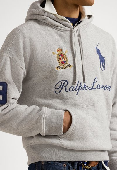 Mężczyzna w jasnoszarej bluzie z kapturem Ralph Lauren z haftowanymi logo, herbem i numerem 3 na rękawie, ręka w przedniej kieszeni.