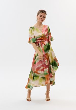Kobieta stojąca w kolorowej, zwiewnej sukience kopertowej z zielonymi, pomarańczowymi i różowymi wzorami akwarelowymi oraz beżowych sandałach na obcasie z paskami.