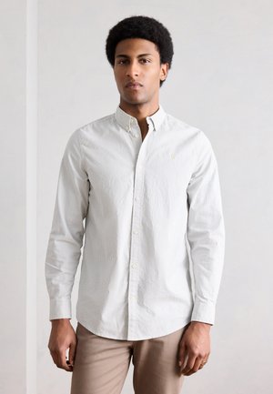 Uomo che indossa una camicia a righe grigio chiaro con bottoni e pantaloni beige, in piedi davanti a uno sfondo chiaro e uniforme, rivolto in avanti.