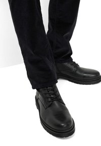 Zwarte leren veterschoenen met een gladde afwerking, stevige zool en subtiele stikdetails. Corduroy broeken in donker marineblauw zijn gedeeltelijk zichtbaar.