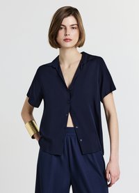 Chemise bleu marine à manches courtes avec boutons sur le devant et col, confectionnée dans un tissu léger. Portée avec un pantalon assorti et un bracelet manchette doré.