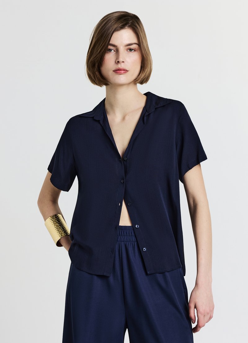 Chemise bleu marine à manches courtes avec boutons sur le devant et col, confectionnée dans un tissu léger. Portée avec un pantalon assorti et un bracelet manchette doré.